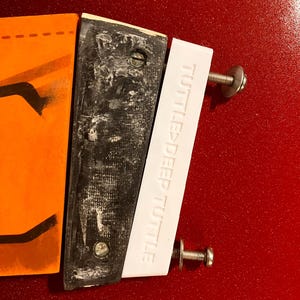 Pode incluir: Close-up de um objeto desmontado com uma seção laranja, uma seção preta e uma seção branca com o texto "TUTTLE DEEP TUTTLE". O objeto é mantido junto com parafusos e está sobre uma superfície vermelha.