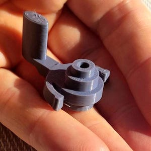 May include: A small, grey, 3D-printed plastic component featuring a central cylindrical base with a tiered, stepped design, a hollow circular opening at the top, and an L-shaped arm extending from the side with a curved, hook-like attachment.