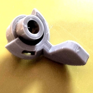 Peut inclure: Un composant en plastique gris clair avec une ouverture circulaire et une extension en forme de levier. L'objet semble être une petite partie d'un mécanisme plus grand, possiblement pour contrôler le flux ou le mouvement. Design simple et fonctionnel.