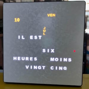 Peut inclure: Une horloge à mots numérique carrée avec une surface grise texturée. L'écran affiche le texte IL EST SIX HEURES MOINS VINGT CINQ en lettres blanches, la date 10 VEN JUL en jaune, et un petit voyant lumineux rouge sur le côté droit.