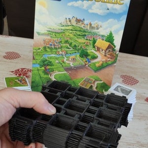 Puede incluir: Un organizador de fichas de plástico negro para el juego de mesa Carcassonne. El organizador está hecho de varias capas apiladas con ranuras para las fichas del juego. La caja del juego está en el fondo con el título del juego "Carcassonne" impreso en ella.