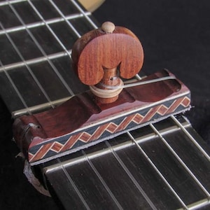 May include: A wooden guitar capo with a decorative design. The capo is made of dark brown wood with a lighter wood accent and a patterned inlay of brown, black, and white. It is placed on the fretboard of a guitar.