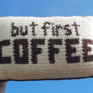 Op de afbeelding: Een wit gebreid kussen met bruine gebreide letters die "but first coffee" lezen.