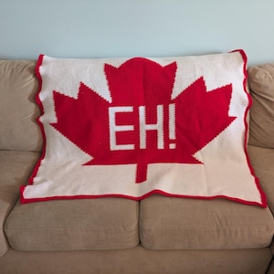 Könnte beinhalten: Ein weißes und rotes gehäkeltes Kissen mit einem großen Ahornblatt-Design. Das Ahornblatt ist rot mit dem Wort "EH!" in Weiß. Das Kissen hat einen gewellten roten Rand und wird auf einem beigen Sofa präsentiert.