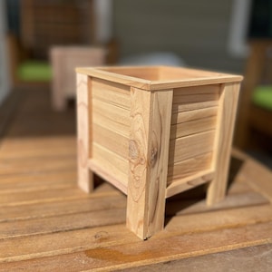 May include: A square wooden planter box with a natural finish. The box is constructed with horizontal slats and supported by four sturdy legs. The planter is designed for outdoor use and is sitting on a wooden surface.