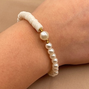 Könnte beinhalten: Ein weißes Perlenarmband mit goldenen Akzenten. Das Armband besteht aus kleinen, runden Perlen und hat ein schlichtes, elegantes Design.
