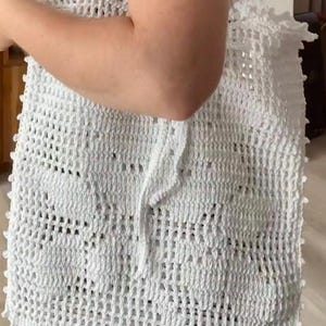 Könnte beinhalten: Eine weiße Häkeltasche mit einem durchbrochenen Netzmuster und einem sich wiederholenden Blumenmotiv. Die Tasche verfügt über zwei passende weiße Häkel-Schulterriemen und eine dekorative Picot-Kante an den Seiten sowie an der oberen Öffnung.