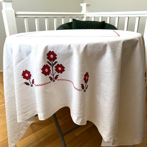 May include: White round tablecloth with red floral embroidery. The embroidered flowers are clustered in groups of three, with a connecting red line. The tablecloth is draped over a round table with a gray metal frame.