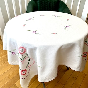 May include: White round tablecloth with embroidered pink and purple flowers. The tablecloth is on a round table with a black metal base.