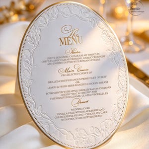May include: An oval-shaped white menu card with gold foil lettering and an embossed rose border. The text lists a starter of Caesar salad, main courses of beef or chicken with sides, and dessert options of vanilla or chocolate wedding cake.
