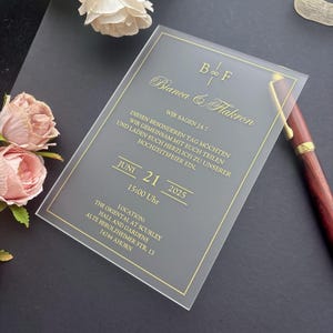 May include: A rectangular, frosted acrylic wedding invitation featuring gold foil text and a thin gold border. The invitation displays the names Bianca and Flakron, the date June 21, 2025, and event details in German, including the location in Ahorn.