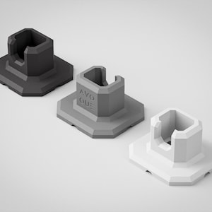 Puede incluir: Tres soportes geométricos angulares en tonos grises. Los soportes tienen una base cuadrada y una estructura central con la parte superior abierta. El soporte central tiene el texto "AVO QUE" en un lateral. Los soportes están sobre un fondo blanco.