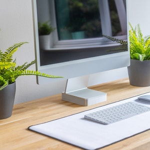 Puede incluir: Una moderna configuración de computadora en un escritorio de madera clara. El conjunto incluye un monitor de computadora plateado, teclado y ratón sobre una alfombrilla blanca. Dos plantas verdes en macetas flanquean el monitor. El fondo es una pared blanca y una ventana.