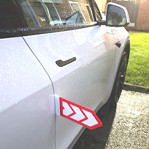 Pode incluir: Carro branco com um sinal em forma de seta vermelha e branca fixado na lateral. O sinal indica a direção. O carro está estacionado numa superfície de betão junto a uma área relvada.