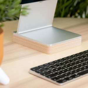 May include: A silver metal tablet or phone stand featuring a base constructed from multiple stacked layers of light-colored plywood. The stand has a smooth, angled support surface and a flat, rectangular base with visible wood grain edges.