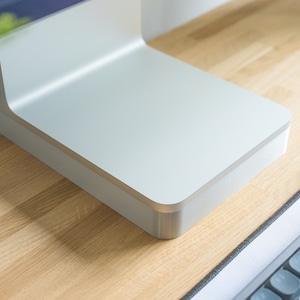 May include: A silver-colored metal computer monitor stand with a flat, rectangular base and a vertical support arm. The stand features a smooth, matte finish and rounded corners, designed to elevate a display screen on a desk surface.