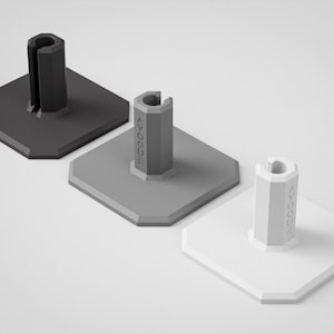 May include: Three geometric, minimalist stands in shades of gray. Each stand has a square base and a cylindrical top with a curved slot. The middle stand has the word "AVOGUE" printed on the side.