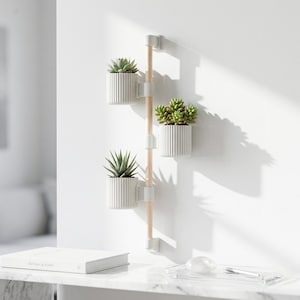 May include: A wall-mounted planter featuring three white, ribbed pots, each containing a succulent plant. The pots are secured to a wooden pole with white brackets. The design is set against a white wall, offering a minimalist style.