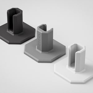 Pode incluir: Três suportes geométricos com base hexagonal em tons de cinza. Cada suporte apresenta um suporte vertical em forma de U. Os suportes estão dispostos sobre uma superfície branca. A palavra "AVOCUE" está impressa no suporte do meio.