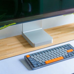May include: A gray and orange mechanical keyboard sits on a white mousepad in front of a computer monitor. The keyboard has gray keycaps with orange accents. The monitor is silver and black, and the desk is wood.