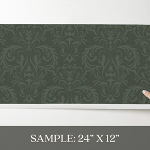 The product is a 24 by 12 inch rectangular wallpaper sample made from paper-based material. It has a dark green matte finish with a tone-on-tone damask pattern in a slightly darker shade. The pattern forms a repeating arrangement of symmetrical, ornate motifs. The surface shows a subtle, raised texture along the pattern lines.