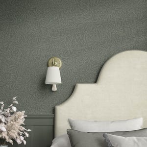 May include: A bedroom scene featuring a patterned wallpaper in shades of green and gray. A cream-colored headboard and pillows are visible. A gold-toned wall sconce with a white lampshade and tassel hangs on the wall. Decorative dried flowers are in a vase.