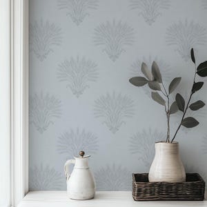 May include: A light blue wall with a repeating floral pattern. A white pitcher with a wooden stopper and a cream-colored vase with dark green leaves sit on a white surface. A dark brown wicker basket is also present.