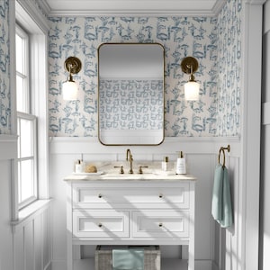 May include: A white bathroom vanity with a marble countertop, brass fixtures, and a rectangular mirror. The walls are decorated with a blue and white fish pattern wallpaper. Two sconces with white glass shades flank the mirror. A light blue towel hangs on a brass towel ring.