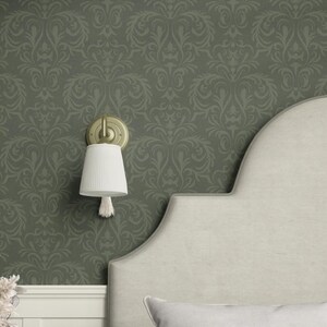 This wall-mounted sconce features a textured cream-colored fabric shade in a cylindrical form tapering subtly toward its open base. A single tassel extends from the shade’s lower center. The supporting arm and wall plate are metal finished in a brushed brass-like tone.
