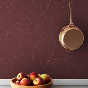 Op de afbeelding: Een koperen steelpan hangt aan een bordeauxrode muur met een bloemenpatroon. Een houten kom gevuld met rode appels staat op een wit oppervlak. De scène roept een rustieke keukenesthetiek op.