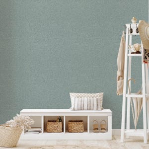 Op de afbeelding: Een serene interieurscène met een lichtblauwe muur met een subtiel bladerpatroon. Een witte bank met geweven manden en een kussen met patroon staat eronder. Een witte kapstok met een badjas, hoed en paraplu, creëert een rustgevende, neutrale esthetiek.