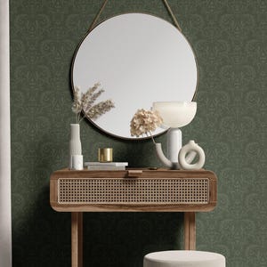 The image shows a round mirror with a slender metallic frame in a brushed gold finish. Below it sits a rectangular wooden console table with a woven rattan front panel and four slender tapered legs. In front of the table is a cylinder-shaped stool featuring a smooth beige upholstered top and a vertically pleated fabric skirt around its lower half.