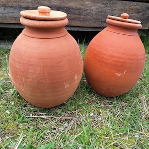 Può includere: Due vasi di terracotta con coperchi. I vasi sono di colore marrone-rossastro e hanno una forma arrotondata. I coperchi sono piatti con un piccolo pomello sulla parte superiore. I vasi sono appoggiati su erba verde davanti a uno sfondo di legno.