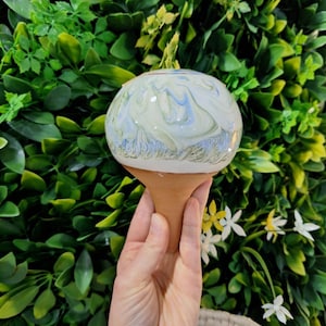 Può includere: Un annaffiatoio decorativo in ceramica con una parte superiore rotonda, marmorizzata bianca, blu e verde e una base in terracotta. L'annaffiatoio è progettato per rilasciare lentamente l'acqua nel terreno, mantenendo le piante idratate.