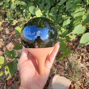 Può includere: Una sfera da giardino nera e sferica con una base in terracotta. La superficie riflettente riflette il cielo e il verde circostante. La sfera è tenuta in mano, con uno sfondo di fogliame verde.