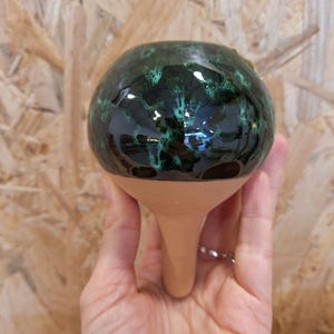 Può includere: Un vaso di fiori in ceramica fatto a mano con una finitura verde scuro lucida e un motivo maculato. Il vaso ha una parte superiore arrotondata e una base lunga e affusolata. Il vaso è tenuto in mano, mostrando la sua forma e il suo design unici.
