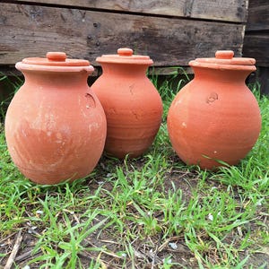 Puede incluir: Tres ollas de terracota con tapas, colocadas sobre hierba verde. Las ollas son de un color marrón anaranjado cálido, con una textura ligeramente rugosa. Las tapas tienen pequeños tiradores redondos. El fondo presenta tablas de madera desgastadas.
