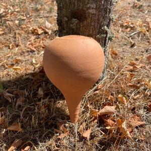 Può includere: Un tappo per bottiglia rotondo in ceramica di colore terracotta con una punta appuntita. Il tappo è appoggiato su un letto di foglie secche ed erba.