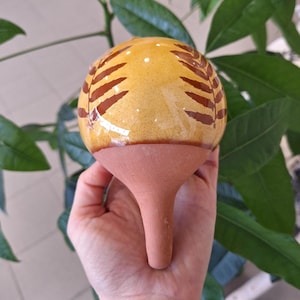 Può includere: Un annaffiatoio in ceramica per piante con una parte superiore sferica gialla decorata con motivi a foglie marroni. L'annaffiatoio ha una base e uno stelo color terracotta. Lo sfondo comprende foglie verdi.