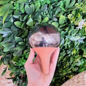 Può includere: Un globo decorativo in vetro con una superficie riflettente a specchio, montato su una base a forma di cono color terracotta. Il globo è tenuto in mano su uno sfondo di fogliame verde.
