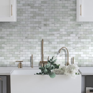 May include: A kitchen scene featuring a white farmhouse sink with a gold faucet and hardware. The backsplash is made of white and gray marble-look tiles. Greenery and white flowers are arranged near the sink, creating a fresh and inviting atmosphere.