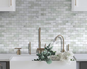 May include: A kitchen scene featuring a white farmhouse sink with a gold faucet and hardware. The backsplash is made of white and gray marble-look tiles. Greenery and white flowers are arranged near the sink, creating a fresh and inviting atmosphere.