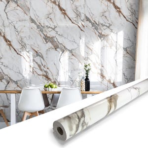 May include: A roll of marble-patterned wallpaper is displayed in front of a dining room with a marble wall. The wallpaper roll is white with gray and brown veining. The dining room has a wooden table, white chairs, and a black vase.
