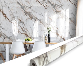 May include: A roll of adhesive contact paper featuring a white marble pattern with grey and brown veining. The roll is positioned in the foreground, while the background displays a wall covered in the same marble pattern behind a dining table.
