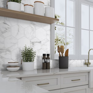 May include: White hexagonal marble tile backsplash in a kitchen with white cabinets, a wooden shelf with white ceramic jars, and a window with a view of trees.