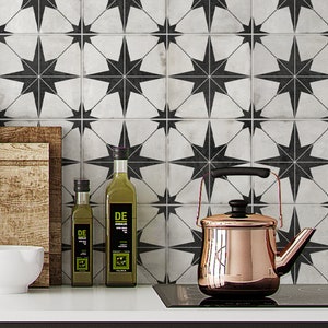 May include: A copper kettle with a black handle sits on a black stovetop in front of a white tiled backsplash with black star designs. Two glass bottles of olive oil with green labels are on a countertop to the left of the kettle. The label on the larger bottle reads "DE ANDALUS Extra Virgin Olive Oil".