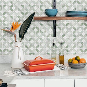 May include: A kitchen scene with a white countertop and a patterned tile backsplash. A bright orange rectangular baking dish sits on a white and black striped towel. A white vase holds a bird of paradise flower and a large green leaf. A bowl of fruit and blue dishes are on a shelf.