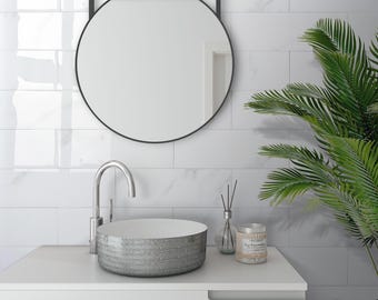 May include: A modern bathroom scene featuring a round black mirror, a silver vessel sink, and a chrome faucet. The sink sits atop a white cabinet, with a candle and diffuser adding to the spa-like ambiance. White marble-look tiles and a green plant complete the decor.