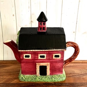 Può includere: Una teiera in ceramica a forma di fienile rosso con tetto nero e torretta. L'oggetto presenta un manico con texture effetto legno marrone, un beccuccio rosso e una base verde testurizzata. Le pareti hanno un motivo a listelli con tre finestre.