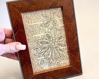 Könnte beinhalten: Ein rechteckiger Bilderrahmen mit dunkelbrauner Holzmaserung und einem seilartig texturierten Innenrand. Der Rahmen enthält ein Blatt Papier im Vintage-Stil, das mit schwarzem Text und einer schwarzen Strichzeichnung eines großen Blumenmusters bedruckt ist.